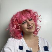 A girl with shoulder length pink curly hair is shown wearing a white t shirt