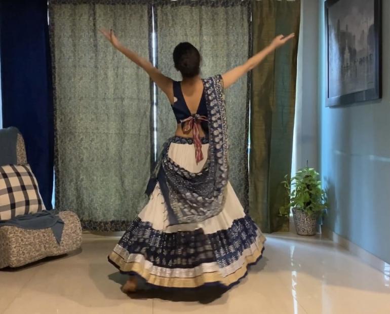 a girl in a long white skirt with indigo and gold borders, wearing a matching indigo blouse. Her hair is tied in a bun. Her back is facing the camera. Her hands are spread up in a pose.