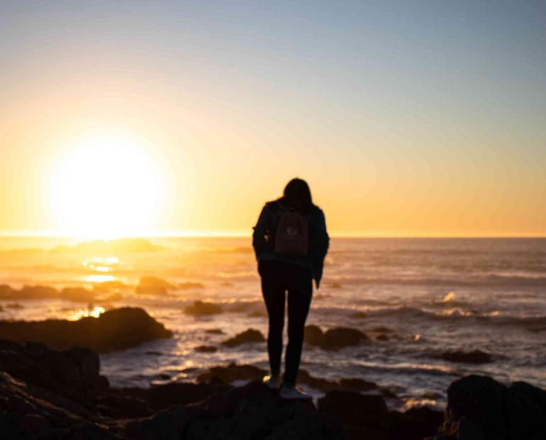 A girl stands in front of the ocean, the sun setting over the horizon.