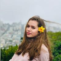 Chica rubia oscuro con flor amarilla en el la oreja, usando un suéter de cuello tortuga rosa pálido y el fondo de la fotografía es la ciudad de San Francisco. 