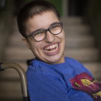 A teenage girl in a wheelchair; smiling brightly, with short hair and wearing a Superman T-shirt.