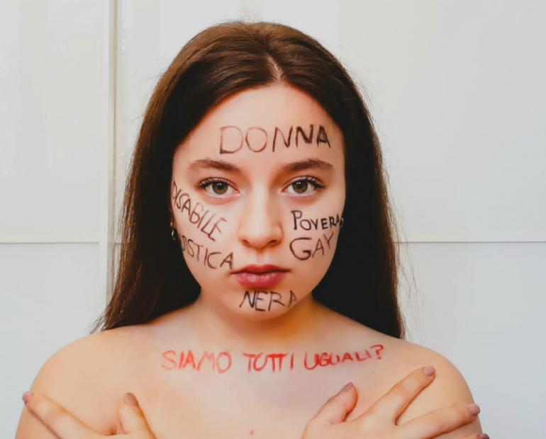 A young adult is posing straight in front of the camera, words such as "poor", "disabled", "gay" and others are written on her face. On her shoulders, you can read: "Are we all really equal?". Every word is written in Italian.