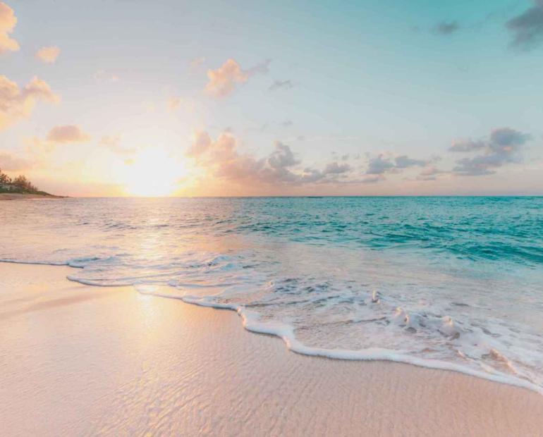 A beach with white sand, cyan water, and the sun setting in the back.