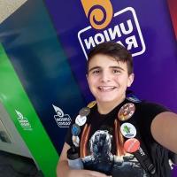 A teenager with brown hair and a black t-shirt standing in front of a wall with multiple colors